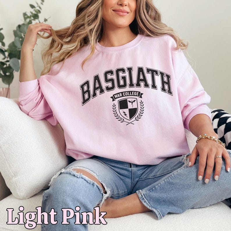 Peut inclure: Sweat-shirt rose clair avec l'inscription "BASGIATH WAR COLLEGE" en noir. Le design comprend un &eacute;cusson avec un bouclier, un oiseau et un &eacute;clair. Les mots "Light Pink" sont &eacute;crits en cursive en bas.