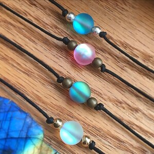 Puede incluir: Primer plano de cuatro collares con cuentas de vidrio iridiscente en tonos azules, rosas y verdes. Las cuentas est&aacute;n ensartadas en cordones negros con detalles plateados y dorados. El fondo es una superficie de madera.