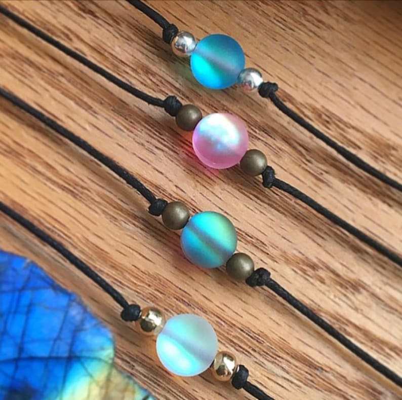 Puede incluir: Primer plano de cuatro collares con cuentas de vidrio iridiscente en tonos azules, rosas y verdes. Las cuentas est&aacute;n ensartadas en cordones negros con detalles plateados y dorados. El fondo es una superficie de madera.