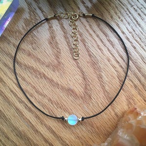 Puede incluir: Un delicado collar gargantilla con una peque&ntilde;a cuenta iridiscente en el centro. La cuenta est&aacute; suspendida de un fino cord&oacute;n negro con detalles met&aacute;licos dorados y un cierre ajustable. El collar se exhibe sobre una superficie de madera.