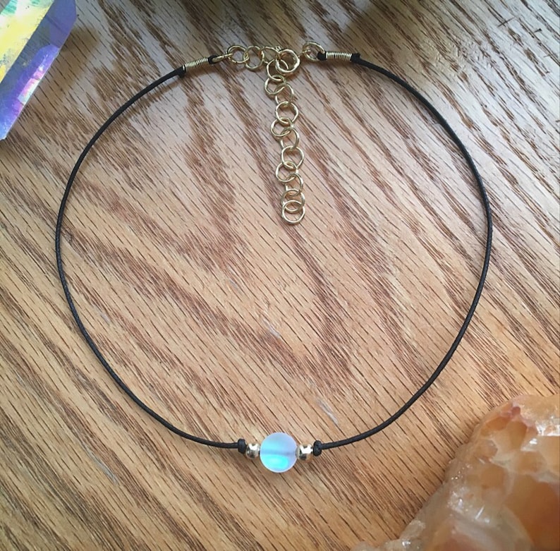 Puede incluir: Un delicado collar gargantilla con una peque&ntilde;a cuenta iridiscente en el centro. La cuenta est&aacute; suspendida de un fino cord&oacute;n negro con detalles met&aacute;licos dorados y un cierre ajustable. El collar se exhibe sobre una superficie de madera.