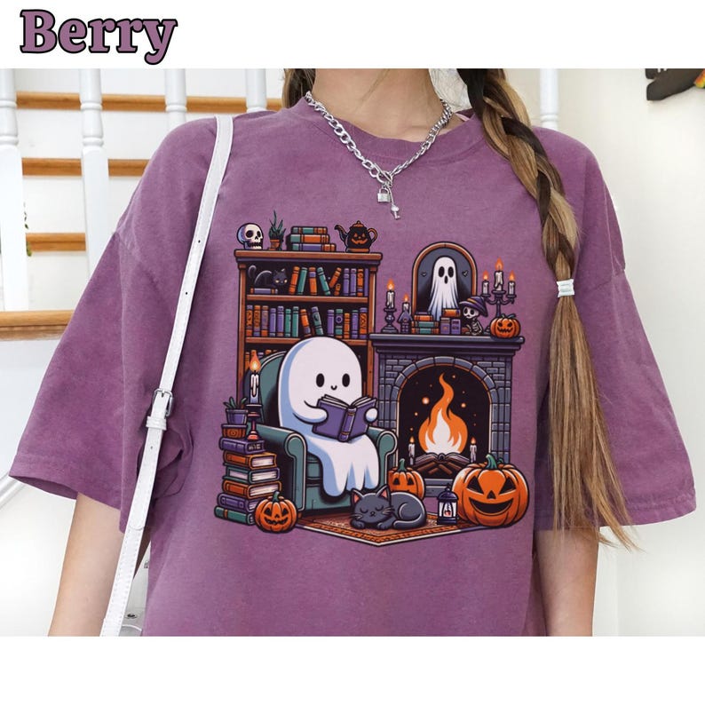 May include: Berry-colored t-shirt featuring a cartoon ghost reading a book in a cozy library setting. The design includes a fireplace, pumpkins, a black cat, and bookshelves. The word "Berry" is printed at the top.