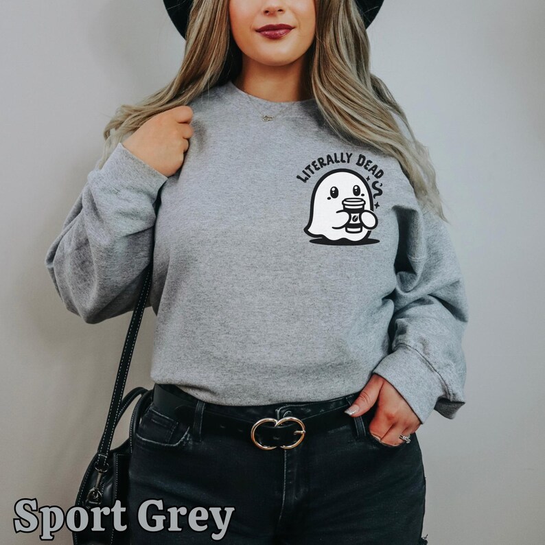 Pu&ograve; includere: Una felpa grigia con un fantasma che tiene una tazza di caff&egrave; e la scritta "Literally Dead" stampata sul davanti. La modella indossa jeans neri e una cintura nera.