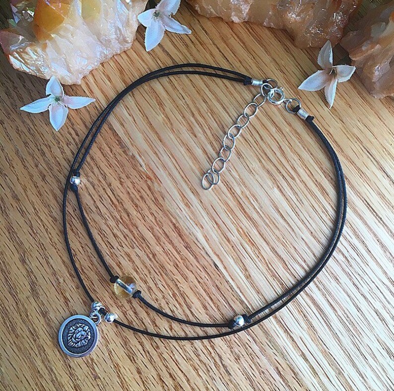 May include: A black cord choker necklace with a silver lion charm and a small amber bead. The necklace has a silver clasp.