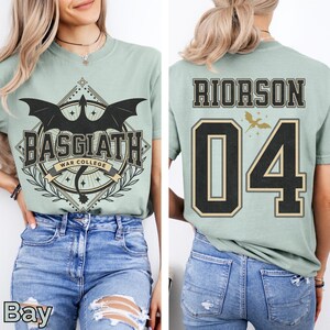 K&ouml;nnte beinhalten: Hellgr&uuml;nes T-Shirt mit dem Text "BASGIATH WAR COLLEGE" und einem Drachenmotiv auf der Vorderseite. Auf der R&uuml;ckseite des Shirts steht "RIORSON 04" in Schwarz und Gold. Das Shirt besteht aus einem weichen Material.
