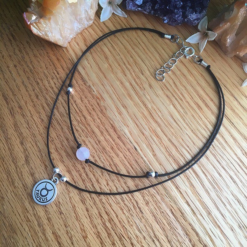 May include: A black cord necklace with a silver Taurus charm and a pink bead. The necklace is layered and has silver beads.