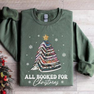 Könnte beinhalten: Ein grünes Sweatshirt mit einem Weihnachtsbaum aus Büchern. Der Baum ist mit Lichtern und einem Stern an der Spitze geschmückt. Der Text "All Booked for Christmas" ist unter dem Baum gedruckt.