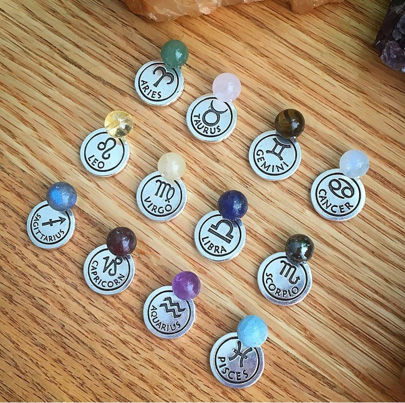 May include: Twelve silver charms with zodiac symbols and corresponding gemstones. The charms are arranged in a circle on a wooden surface.