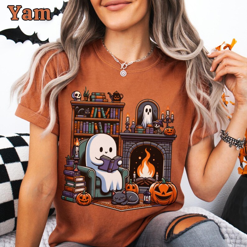 May include: A burnt orange t-shirt featuring a Halloween-themed graphic. The design depicts a ghost reading in a cozy room with a fireplace, bookshelves, pumpkins, and a black cat. The word "Yam" is in the top left corner.