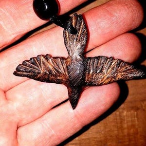 May include: A wooden bird pendant with intricate details, carved in a dark brown colour. The pendant is attached to a black cord.