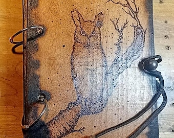 Puede incluir: Diario de madera con un diseño de búho pirograbado. La cubierta presenta una ilustración detallada de un búho posado en una rama, con un cierre de cordón marrón oscuro y anillas metálicas. La madera tiene un borde envejecido y quemado.