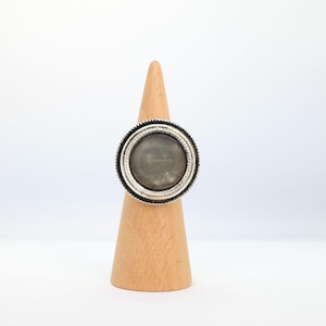 Könnte beinhalten: Ein silberner Ring mit einem großen, runden, grauen Stein in der Mitte. Der Ring hat einen strukturierten Rand und wird auf einem hölzernen Ringhalter präsentiert. Das Design ist schlicht, aber elegant und eignet sich für den Alltag oder besondere Anlässe.