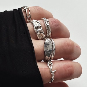 Peut inclure: Un ensemble de quatre bagues en argent avec des designs uniques. Les bagues sont empilées sur un doigt et présentent une variété de textures et de formes, notamment un design en forme de brindille, un ovale lisse et deux bagues avec une texture rugueuse et organique.