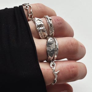 May include: A hand displays several silver rings with unique, textured designs. The rings vary in shape, including organic, branch-like, and hammered metal styles. The rings are worn on multiple fingers, showcasing the different designs.