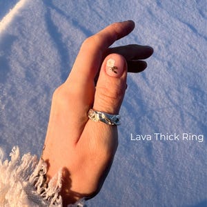 A close-up of a left hand in natural winter sunlight, resting against textured snow. The thumb wears a bold Lava Thick Ring with a raw, molten silver texture and organic, sculptural surface. A subtle floral nail design is visible on the thumbnail. The image includes a label identifying the ring. The overall aesthetic feels earthy, elemental, and expressive — contrasting warm skin tones with the cool, icy background.