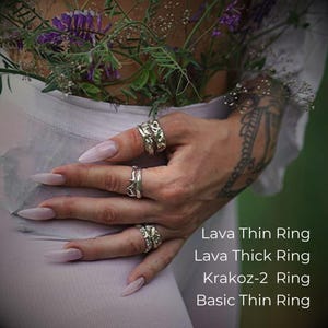 Close-up of a tattooed hand adorned with four handcrafted silver rings in various textures and widths, resting on the hip of a person dressed in sheer white fabric. Wildflowers are tucked into the waistband, adding a natural, romantic touch. The rings shown are labeled: Lava Thin Ring, Lava Thick Ring, Krakоz-2 Ring, and Basic Thin Ring — all worn together to create a bold, stacked look.