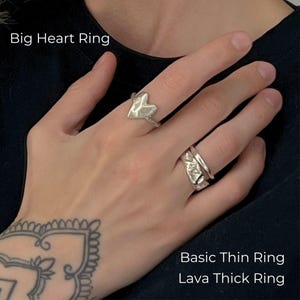 A close-up of a tattooed hand wearing three silver rings in a modern, artistic style: a bold Big Heart Ring on the index finger, and a stacked combination of a sleek Basic Thin Ring and a textured Lava Thick Ring on the ring finger. The overall look is edgy yet elegant, with a handcrafted, minimalist vibe.