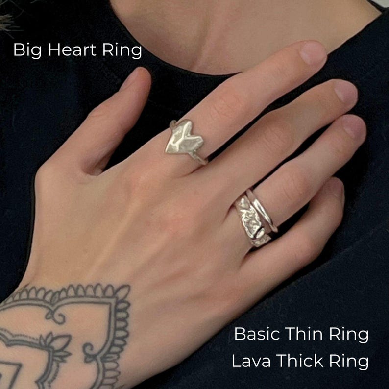 A close-up of a tattooed hand wearing three silver rings in a modern, artistic style: a bold Big Heart Ring on the index finger, and a stacked combination of a sleek Basic Thin Ring and a textured Lava Thick Ring on the ring finger. The overall look is edgy yet elegant, with a handcrafted, minimalist vibe.