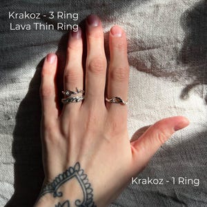 A tattooed hand resting on textured fabric, adorned with three silver rings featuring organic, molten-like designs. Sunlight casts soft shadows, highlighting the intricate details of the jewelry. The rings are labeled Krakoz 1, Krakoz 3, and Lava Thin.