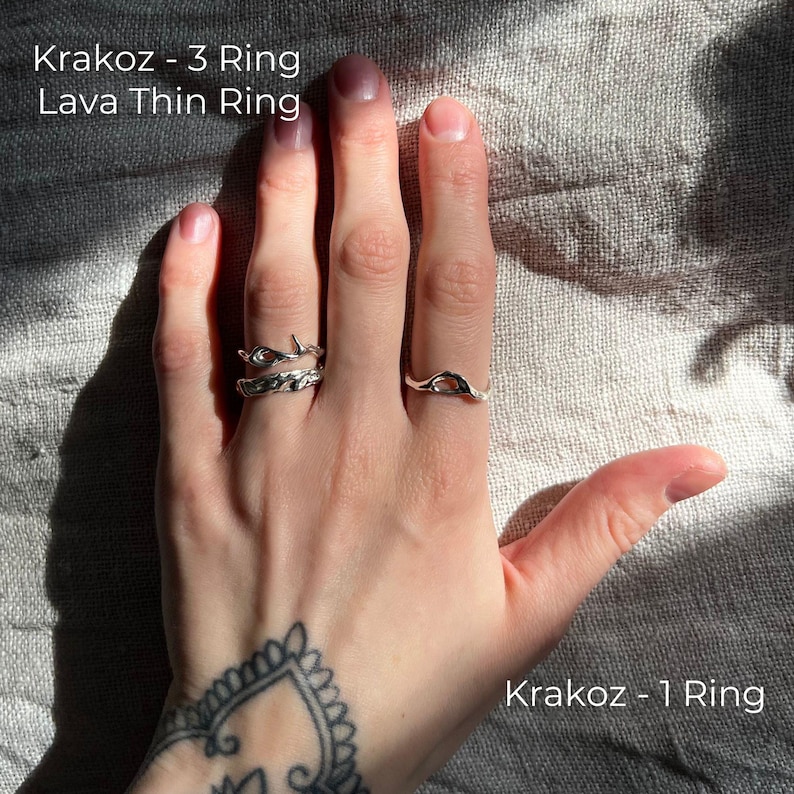 A tattooed hand resting on textured fabric, adorned with three silver rings featuring organic, molten-like designs. Sunlight casts soft shadows, highlighting the intricate details of the jewelry. The rings are labeled Krakoz 1, Krakoz 3, and Lava Thin.