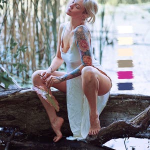 May include: A person in a white dress sits on a log near water, with eyes closed and head tilted back. The person has a floral tattoo on their arm. The background includes water and tall plants.