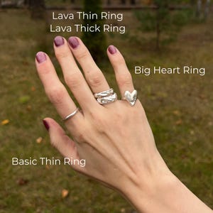 A close-up of a right hand outdoors against a soft-focus grassy background, wearing four artisan silver rings. The ring finger features a stacked pair: Lava Thin and Lava Thick Rings, both with rich, molten textures. The index finger is adorned with a minimalist Basic Thin Ring, while the pinky showcases the expressive, handcrafted Big Heart Ring. Each ring is labeled in the image. The overall aesthetic is earthy, modern, and artistic — a natural fusion of clean form and raw organic detail.