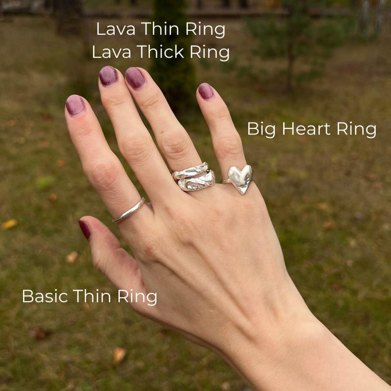 A close-up of a right hand outdoors against a soft-focus grassy background, wearing four artisan silver rings. The ring finger features a stacked pair: Lava Thin and Lava Thick Rings, both with rich, molten textures. The index finger is adorned with a minimalist Basic Thin Ring, while the pinky showcases the expressive, handcrafted Big Heart Ring. Each ring is labeled in the image. The overall aesthetic is earthy, modern, and artistic — a natural fusion of clean form and raw organic detail.