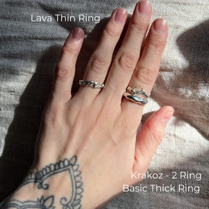 A tattooed hand resting on textured fabric, adorned with three distinct silver rings. The Lava Thin ring features a fluid, molten-like texture, evoking the movement of liquid metal. Krakoz-3 showcases an organic, branch-inspired design with intertwining elements. Basic Thick has a bold, smooth, and slightly irregular shape, resembling a sculpted piece of silver. Sunlight enhances the reflective surfaces of the rings, highlighting their unique craftsmanship.
