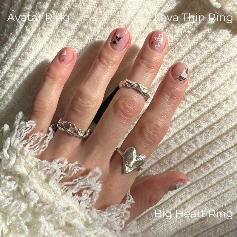 Left hand resting on a soft white textured fabric, styled with three sterling silver rings. On the ring finger, the Avatar Ring features an abstract hollow, molten openwork band with a sculptural, organic silver form. On the middle finger, the Lava Thin Ring shows a fluid, irregular textured band with a raw molten look. On the index finger, the Big Heart Ring stands out as a bold irregular heart, wabi sabi inspired. Modern brutalist, minimalist stacking, artistic and nature inspired style.