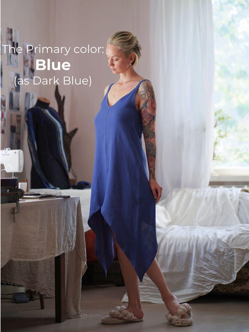 A woman in a blue dress stands in a room with a sewing machine, a table, and a window.