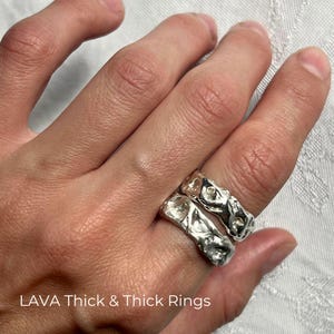 Close-up of a left hand resting on white textured fabric, with two handcrafted solid 925 sterling silver Lava Thick rings stacked on the index finger. The chunky wide bands have uneven wavy edges, sculptural molten relief, organic dents and raised folds, and liquid-like reflections. Their different irregular textures show the character of handmade casting while demonstrating how two Thick variations create a bold freeform statement stack. Overlay text reads “LAVA Thick & Thick Rings.”