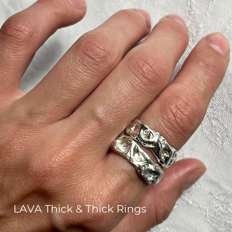 Close-up of a left hand resting on white textured fabric, with two handcrafted solid 925 sterling silver Lava Thick rings stacked on the index finger. The chunky wide bands have uneven wavy edges, sculptural molten relief, organic dents and raised folds, and liquid-like reflections. Their different irregular textures show the character of handmade casting while demonstrating how two Thick variations create a bold freeform statement stack. Overlay text reads “LAVA Thick & Thick Rings.”