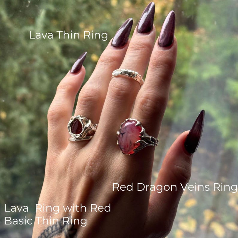 Left hand with long almond-shaped burgundy nails wearing sculptural silver rings. The pointer finger features the Red Dragon Veins Ring with a pink‑red fantasy stone in molten, dragon-inspired claws. A slender Lava Thin Ring sits on the middle finger, while the ring finger shows a stacked Lava Ring with Red and a Basic Thin Ring. The look is edgy, mystical, and artisan, with molten, wabi-sabi textures and indie statement style.