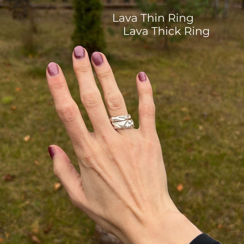 Right hand with short glossy mauve manicure shown against a forest background. On the ring finger are two sterling silver rings: the chunky Lava Thick Ring and the slender Lava Thin Ring. Both have a molten, organic texture with a sculptural, raw aesthetic. The natural outdoor setting enhances the earthy, nature-inspired style of the handmade jewelry.