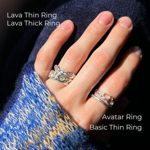 A close-up of a hand wearing four silver rings in sunlight. The ring finger has two stacked rings: Lava Thin Ring and Lava Thick Ring with a molten texture. The index finger features the Avatar Ring with an organic openwork design and the Basic Thin Ring with a smooth finish. The hand rests against a dark blue textured fabric and a cozy sweater in blue, white, and sunflower yellow tones.