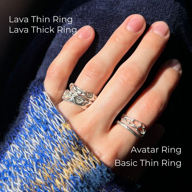 A close-up of a hand wearing four silver rings in sunlight. The ring finger has two stacked rings: Lava Thin Ring and Lava Thick Ring with a molten texture. The index finger features the Avatar Ring with an organic openwork design and the Basic Thin Ring with a smooth finish. The hand rests against a dark blue textured fabric and a cozy sweater in blue, white, and sunflower yellow tones.