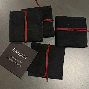 Four handmade rings are carefully wrapped in small black fabric pouches, each secured with a thin red cord. The pouches are arranged on a neutral surface, creating a minimal and slightly moody composition. Next to them lies a dark card with the inscription “EMLAN – your talisman of primal source,” adding a sense of identity and intention to the packaging. The overall presentation feels intimate, tactile, and thoughtfully crafted.