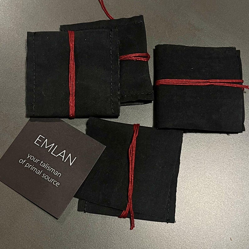 Four handmade rings are carefully wrapped in small black fabric pouches, each secured with a thin red cord. The pouches are arranged on a neutral surface, creating a minimal and slightly moody composition. Next to them lies a dark card with the inscription “EMLAN – your talisman of primal source,” adding a sense of identity and intention to the packaging. The overall presentation feels intimate, tactile, and thoughtfully crafted.