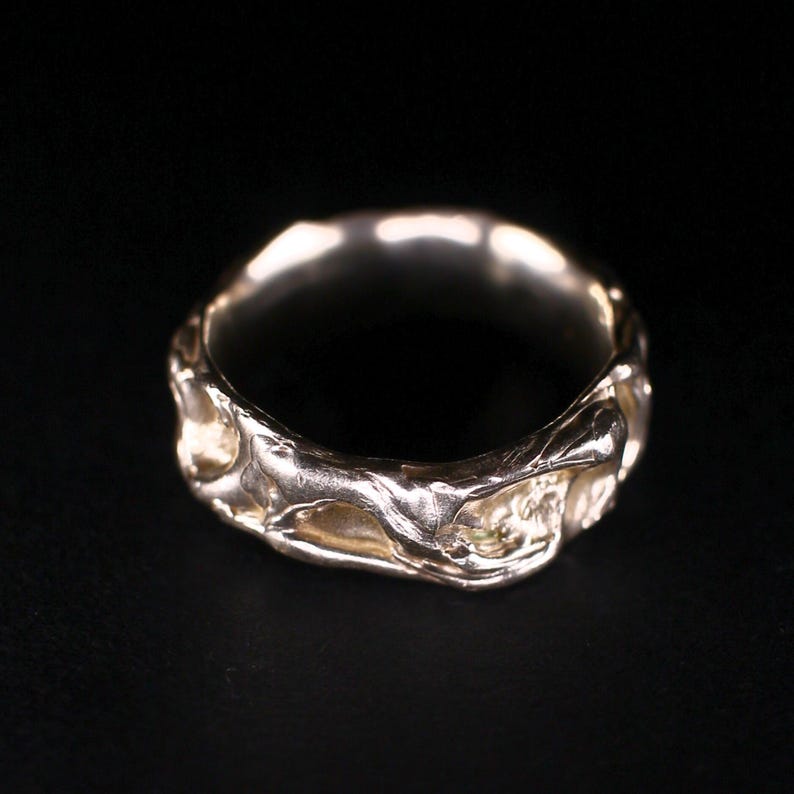 A sterling silver ring is shown close up on a dark background, with a wavy wide band shaped like cooled lava. Its lava uneven ring surface has raw texture, soft molten ridges, and organic hollows that catch the light. This quirky molten ring feels nature inspired, boho wabi-sabi, and bold artsy, with an abstract ebb and flow character for chunky jewelry lovers.