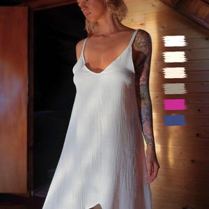 May include: A white, sleeveless dress with a V-neck and spaghetti straps. The dress has a flowing, asymmetrical hemline and a textured fabric. The image is taken indoors with natural light, highlighting the dress's simple elegance.