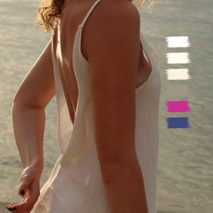 A woman stands by calm sea water at sunset, turning slightly over her shoulder. She wears a white backless dress in soft cotton — an open back slip dress with a low back design and minimalist silhouette. The fabric falls naturally, creating a backless sundress look that feels like beach loungewear, an ibiza resort outfit, or a relaxed bohemian dress for a beach wedding guest. The color palette features White, Milky, Off-White, Beige, Pink, and Blue.