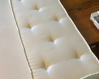 Puede incluir: Cojín rectangular blanquecino con diseño capitoné. Presenta un borde blanco y está colocado sobre una superficie clara. El cojín mide aproximadamente 76 cm de largo y 38 cm de ancho.