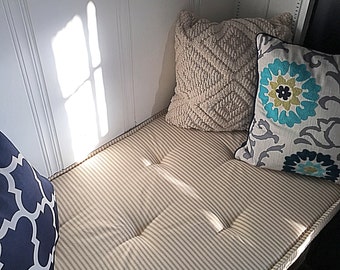 Puede incluir: Un cojín de ventana blanco con tela a rayas y dos almohadas. Una almohada es blanca con un patrón texturizado y la otra es gris con un patrón floral azul y verde.