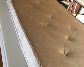 Puede incluir: Un cojín beige capitoné con un borde blanco. El cojín es rectangular y tiene una textura suave y mullida.