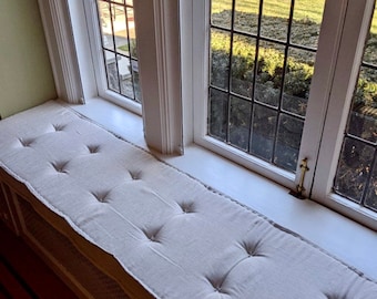 Puede incluir: Un cojín blanco para ventana con capitoné de botones. El cojín está colocado en un asiento de ventana con un marco de ventana pintado de blanco y una vista de un césped verde y árboles en el exterior.
