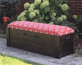 Puede incluir: Un banco de almacenamiento de madera oscura con un cojín rojo y blanco con estampado de flechas. El banco tiene un diseño de flecha de metal en la parte delantera y una manija de metal en el lateral.
