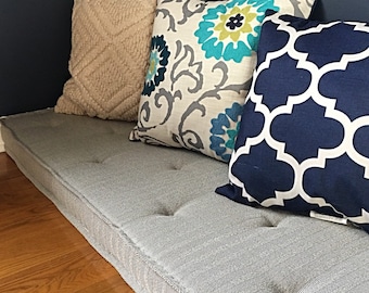 Puede incluir: Un cojín gris acolchado con un patrón geométrico blanco y azul, un patrón floral blanco y gris y una almohada blanca esponjosa. El cojín está colocado en un suelo de madera.