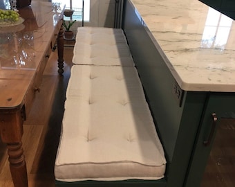 Puede incluir: Un banco integrado con un cojín blanco está unido a una isla de cocina verde oscuro con una encimera de mármol blanco. El banco está frente a una ventana con vista a un jardín.