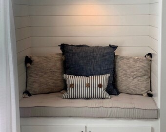 Puede incluir: Un asiento de ventana empotrado blanco con un cojín gris claro y tres almohadas. Las almohadas son de patrón a rayas, azul oscuro sólido y de rayas marrón claro y blanco. Una pared blanca con una lámpara está encima del asiento.