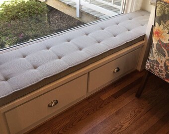 Puede incluir: Un cojín de asiento de ventana blanco con botones capitonnés y una funda de tela gris claro. El cojín está instalado en un asiento de ventana con dos cajones blancos.